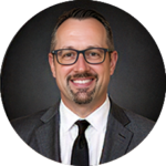 Professional headshot: smiling man with glasses and a goatee, wearing a dark suit and tie, circular crop.