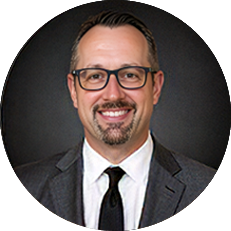 Professional headshot: smiling man with glasses and a goatee, wearing a dark suit and tie, circular crop.