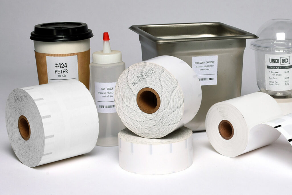 Assorted label rolls and labeling supplies on a white surface, including large white rolls, a glue bottle, and a metal tray.